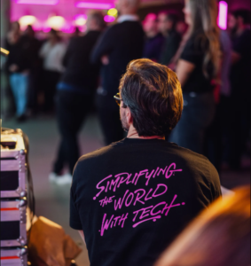 Person auf einer Veranstaltung mit schwarzem T-Shirt und dem pinken Schriftzug ‚Simplifying the world with tech.‘