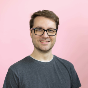 Porträt eines lächelnden Mannes mit kurzen braunen Haaren und schwarzer Brille vor einem rosafarbenen Hintergrund. Er trägt ein dunkelgraues T-Shirt.